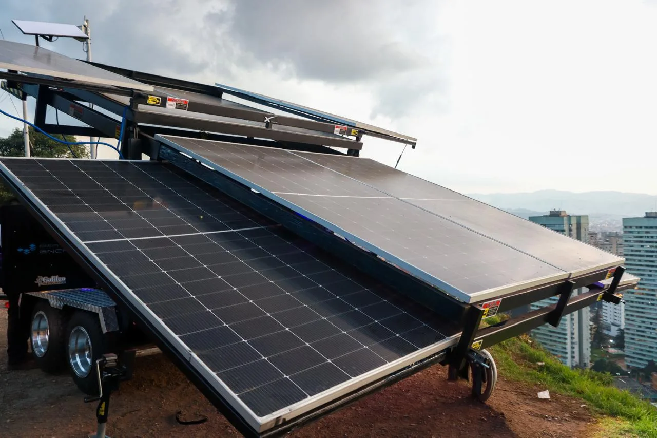 Remolque con paneles solares desplegados en abanico, sobre una loma con edificios de la ciudad al fondo.
